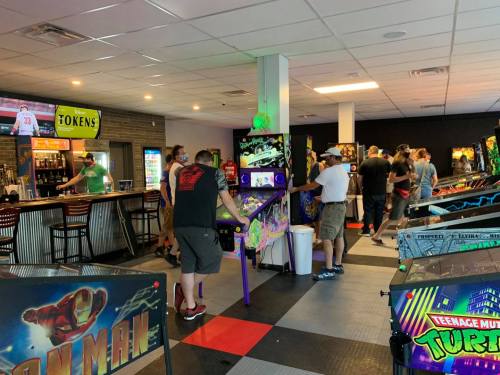 A lively arcade with people playing pinball machines and enjoying drinks at the bar in a vibrant, colorful setting.
