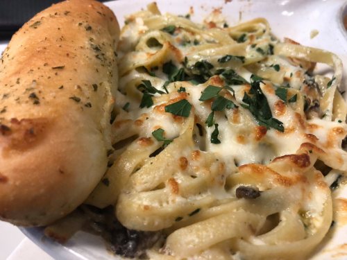 A plate of creamy fettuccine pasta topped with melted cheese and parsley, alongside a garlic breadstick.