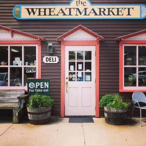 A quaint deli entrance with a pink door, surrounded by potted plants and a sign indicating it's open for take-out.
