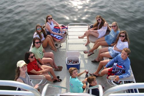 A group of teenagers sitting on a boat, enjoying a sunny day on the water, with drinks and snacks around them.