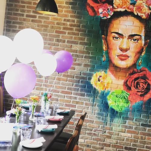 A vibrant dining setup with balloons and flowers, featuring a mural of a woman with flowers in her hair on a brick wall.