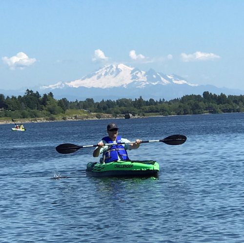 A person kayaking on a calm lake with a snow-capped mountain in the background under a clear blue sky.