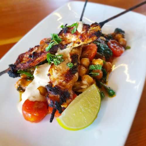 Grilled chicken skewers on a bed of chickpeas and tomatoes, garnished with herbs and a lime wedge.