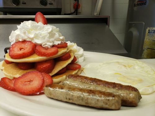 A plate of pancakes topped with strawberries and whipped cream, served with sausage links and fried eggs.