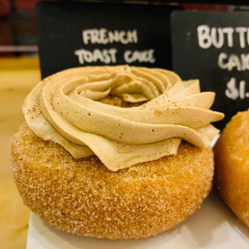 A close-up of a donut topped with swirled brown frosting, labeled "French Toast Cake" on a black sign.