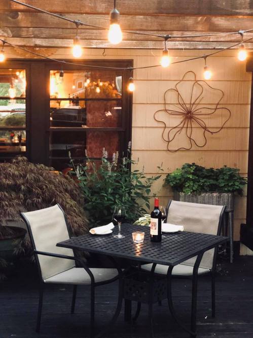 A cozy outdoor dining setup with a table, two chairs, a bottle of wine, and string lights illuminating the space.