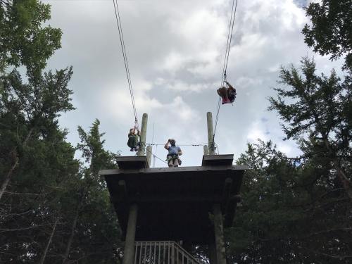 Two people stand on a platform, preparing to zip line through a forested area under a cloudy sky.