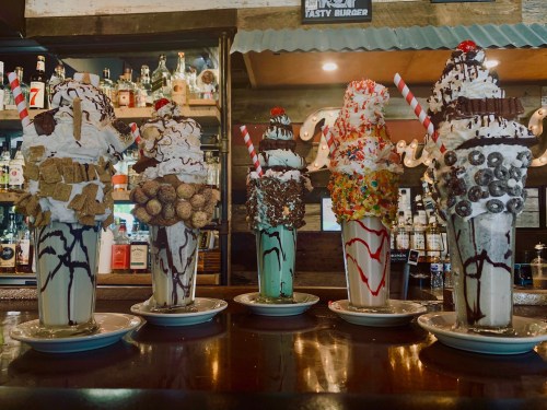 Five extravagant milkshakes topped with whipped cream, candies, and cherries, displayed on a bar counter.