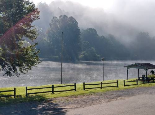 A serene river scene with mist rising, surrounded by trees and a gazebo on the grassy bank.