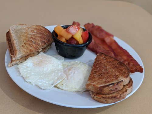 A breakfast plate with two fried eggs, toast, crispy bacon, and a bowl of mixed fruit.