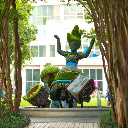 A large topiary sculpture of a dancing figure made of greenery, surrounded by trees and colorful flowers.