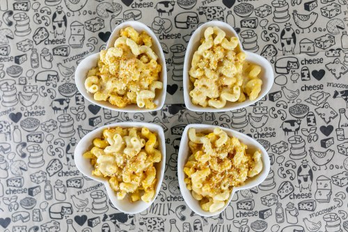 Four heart-shaped bowls of macaroni and cheese on a patterned tablecloth featuring cheese-themed illustrations.