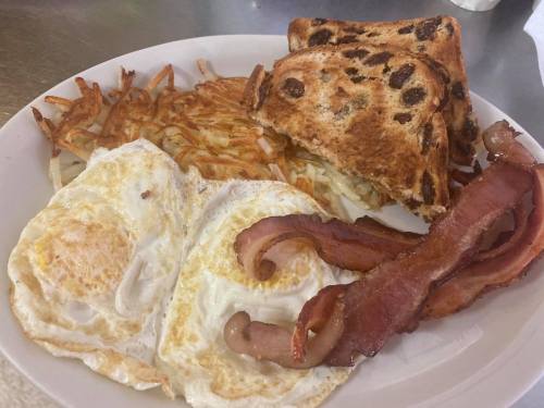 A plate with two fried eggs, crispy bacon, hash browns, and toasted raisin bread.