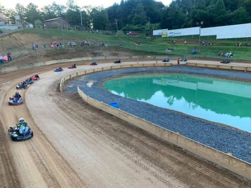 Go-karts race around a curved track beside a pond, with spectators watching from the grassy hillside.