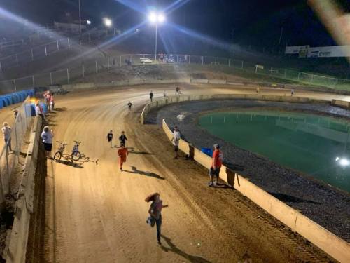 A nighttime view of a dirt racetrack with people running and a bike near a water-filled section. Bright lights illuminate the scene.