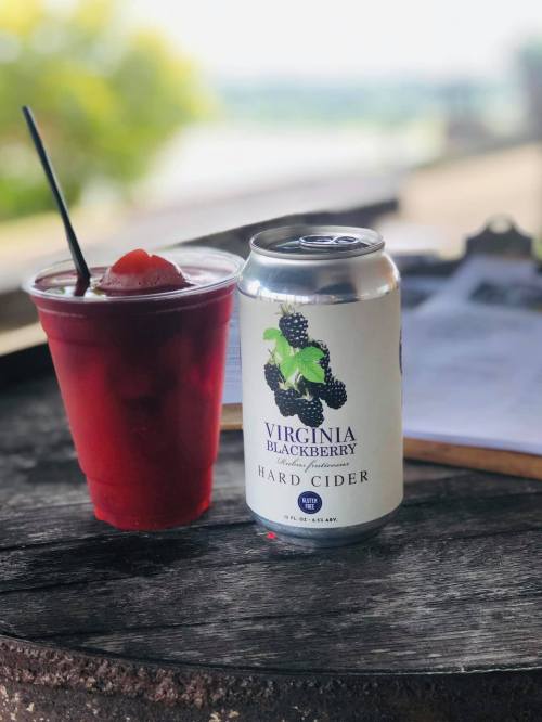 A cup of red drink with a straw next to a can of Virginia Blackberry hard cider on a wooden table.