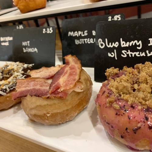Three gourmet donuts on a display: one with chocolate and coconut, one with bacon, and a blueberry donut with streusel.