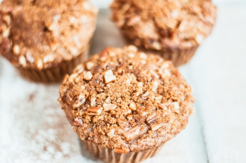 Three freshly baked muffins topped with chopped nuts and a crumbly topping, placed on a light surface.