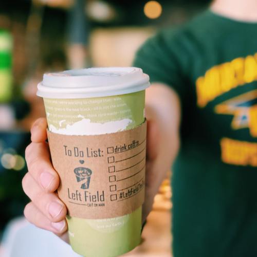 A person holds a coffee cup with a "To Do List" and "Left Field" branding in a cafe setting.