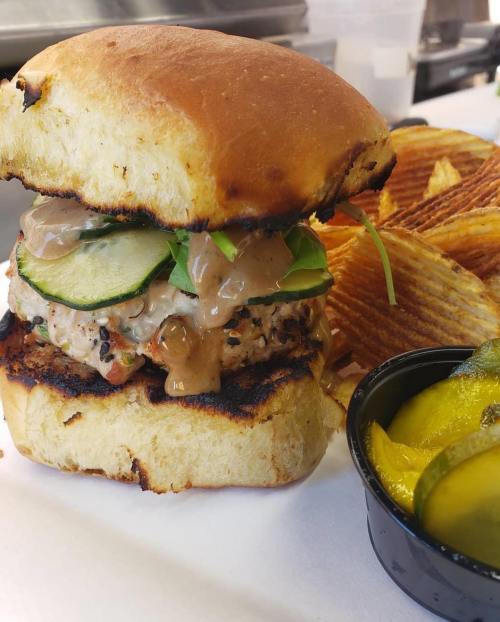 A delicious burger with a grilled patty, fresh greens, cucumber, and sauce, served with crispy chips and pickles.