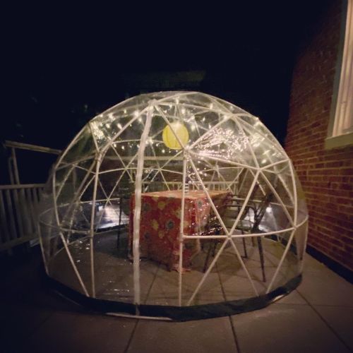 A clear dome-shaped structure with a table inside, illuminated by string lights, set on a patio at night.