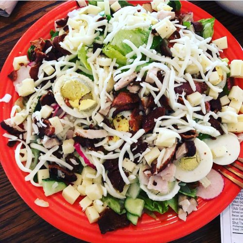A colorful salad with lettuce, cucumbers, bacon, chicken, cheese, and sliced hard-boiled eggs on a red plate.