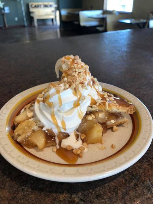 A slice of apple pie topped with whipped cream, caramel drizzle, and chopped nuts on a decorative plate.