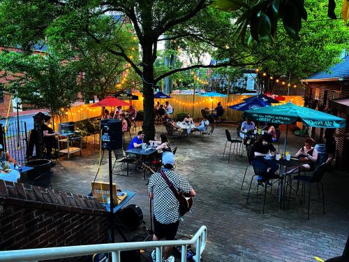 A lively outdoor café scene with people dining, colorful umbrellas, and a musician performing under trees.