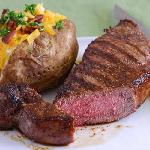 A perfectly cooked steak with a side of a baked potato topped with cheese, bacon, and green onions on a white plate.