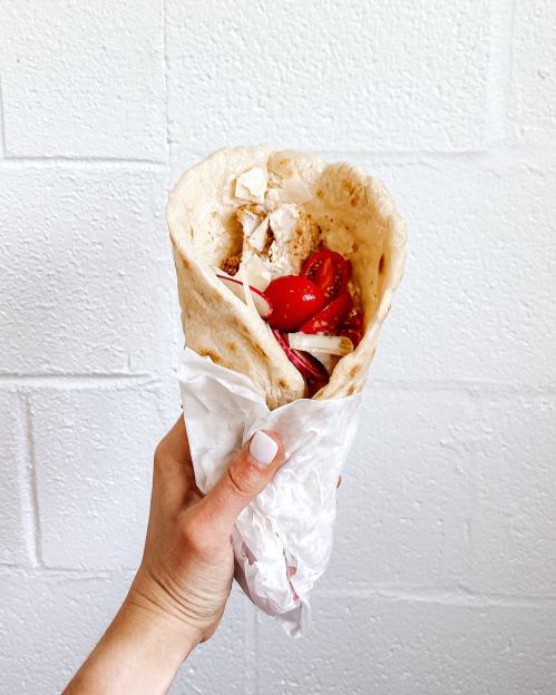 A hand holding a wrap filled with chopped tomatoes and other ingredients, against a white wall.