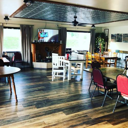 A cozy dining area with wooden floors, tables, chairs, and a TV, featuring large windows and warm lighting.