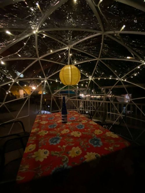 A cozy outdoor dining space with a floral tablecloth, a bottle on the table, and warm lights in a dome structure.