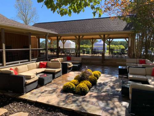 Outdoor seating area with couches, colorful cushions, and flower beds, under a shaded pavilion on a sunny day.