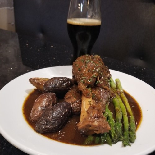 A plate of tender meat with sauce, roasted potatoes, and asparagus, accompanied by a glass of dark beer.