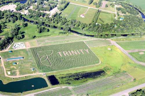 Aerial view of a large corn maze shaped like the word "TRINITY," surrounded by fields and a winding river.