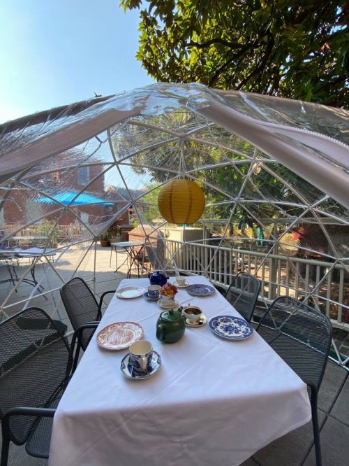 A cozy outdoor dining setup under a clear dome, featuring a table with tea sets and a lantern hanging above.