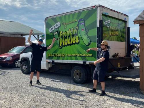 Two people in front of a colorful food truck named "Freakin' Pickles," promoting pickles with a fun design.