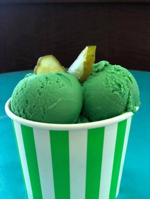 A cup with two green scoops of ice cream topped with pickle slices, set on a turquoise table.