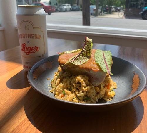 A plate of rice topped with a piece of meat and sauce, alongside a can of Northern Lager, with a sunny outdoor view.