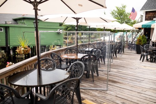 Outdoor dining area with black tables and chairs, umbrellas, and a clear barrier on a wooden deck by the water.