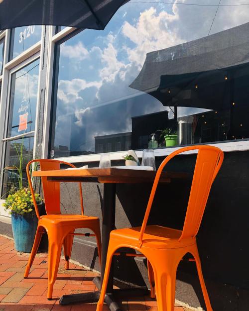 A small outdoor café table with orange chairs, reflecting clouds in the window, surrounded by potted plants.