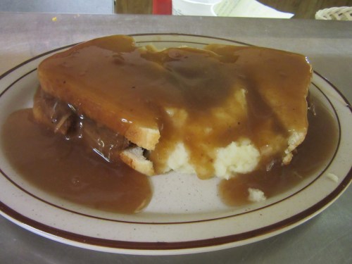A plate with a sandwich topped with gravy, served over mashed potatoes and white bread.