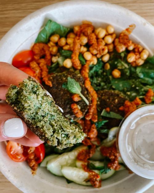 A hand holding a green falafel piece above a bowl filled with salad, chickpeas, and a creamy dressing.
