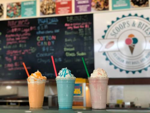 Three colorful milkshakes in tall cups with whipped cream and straws, displayed in front of a menu board.