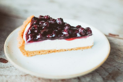 A slice of cheesecake topped with blueberry compote on a white plate, resting on a wooden surface.