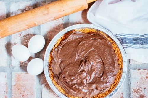 A chocolate pie in a silver tin, surrounded by three eggs and a rolling pin on a textured surface.