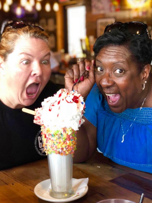 Two excited women with wide smiles pose with a large, colorful ice cream sundae topped with whipped cream and sprinkles.