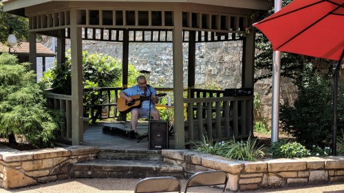 A musician plays guitar in a gazebo surrounded by greenery and stone walls on a sunny day.