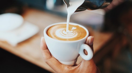 A hand holds a white cup of coffee with a beautiful latte art design as milk is poured on top.