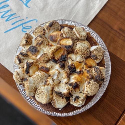 A pie topped with toasted marshmallows, some charred, in a silver pie dish on a wooden table.